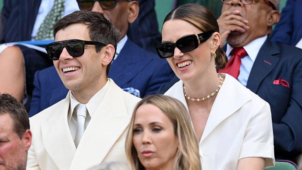 James Righton and Keira Knightley sit side-by-side and smile in the direction of the court. They are both wearing white outfits with black sunglasses.