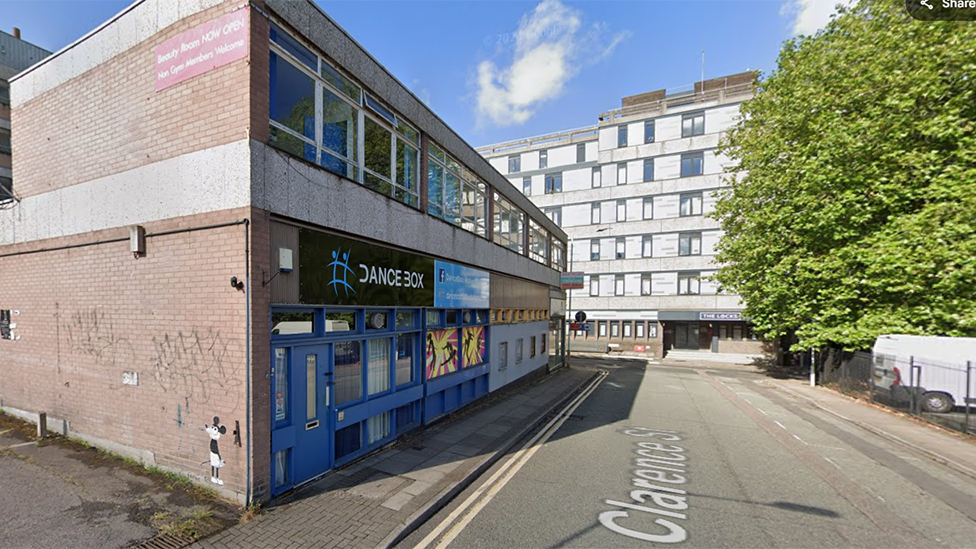 A street view image of a vacant commercial property which has graffiti scrawled on the wall. A large tree is on the right and at the end of the road is a six-storey white building.