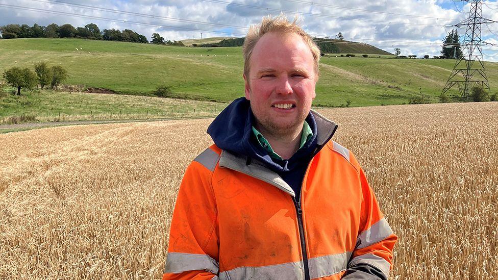 Farmer Cameron Shell, wearing an orange hi-vis coat, standing in a field of barley with green fields beyond and an overheard wire pylon to the right.