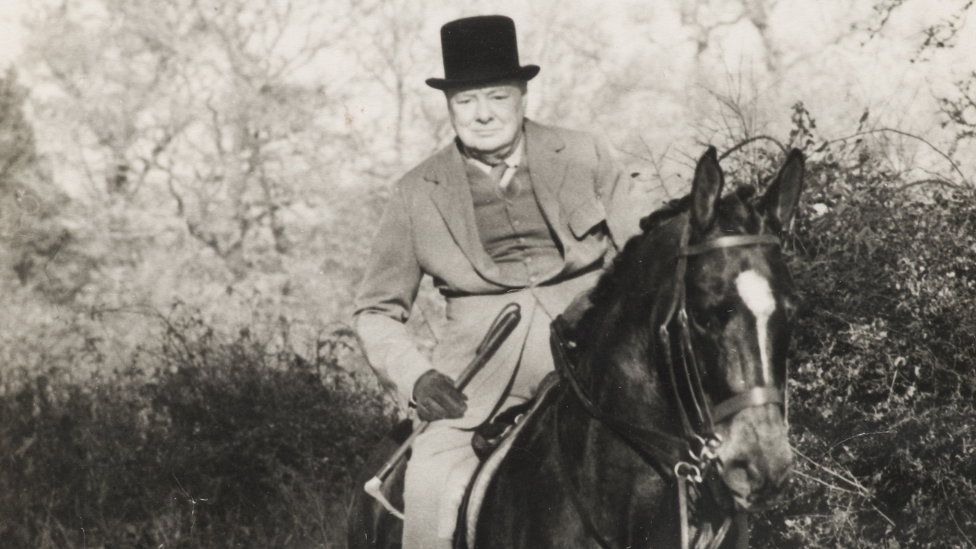 Black and white image of Sir Winston Churchill on horseback. He is well dressed and is wearing his iconic black hat.