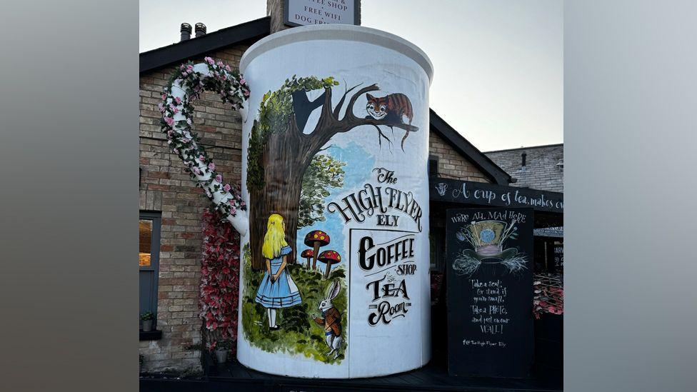 A large mug decoration on the side of a redbrick building. The mug is white with a floral handle and is painted with an image of Alice in Wonderland looking at a tree. There is a grinning cat in the tree. The mug is advertising the name of the pub and coffee shop