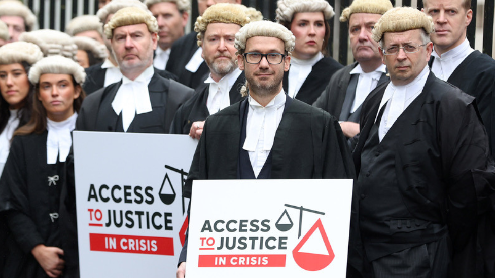 A number of barristers, both male and female, standing in black robes and white wigs, a number of them holding placards saying access top justice in crisis