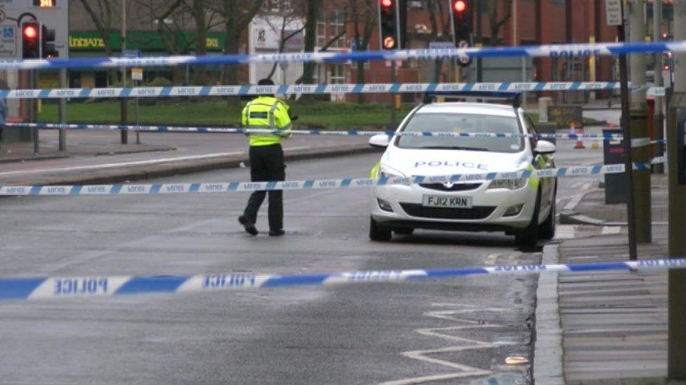 A blue and white police cordon across a road. A police officer in a high visibility jacket can be seen next to a marked police patrol car.