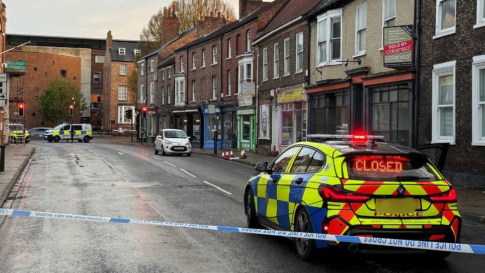 A police car on a city street sits behind a police cordon. Further police vehicles and a parked white car are also visible further down the street.