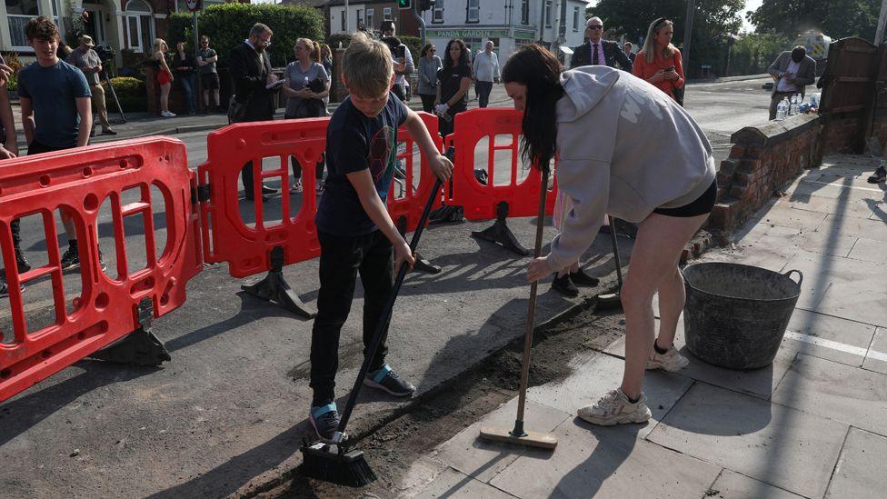 A girl in a grey hoodie and black shorts and a boy in a blue T-shirt and black jeans use brooms to sweep debris off a road in Southport, surrounded by onlookers and orange barriers