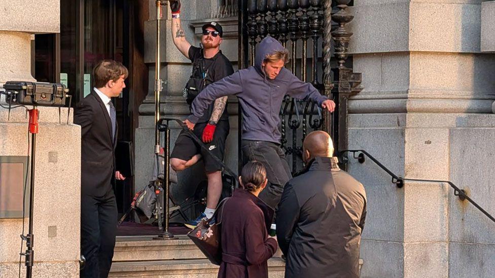 A man is running down the steps of a grey stone building. Past three people. A film crew member in the background is stood in the background.
