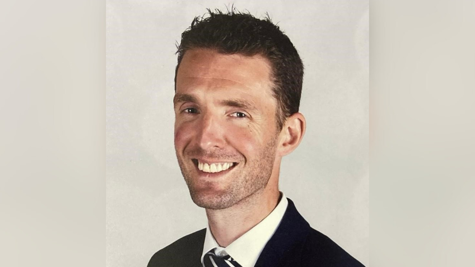 A head shot of Andy burns. He is smiling in front of a beige background, wearing a black suit jacket, white shirt and a black and white striped tie.