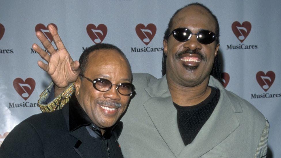 Quincy Jones and Stevie Wonder standing together and smiling on an awards ceremony red carpet in 1999