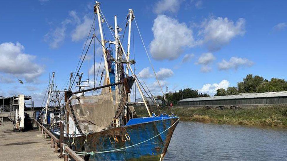 A shrimp fishing boat moored in a dock in King's Lynn. It has a blue body and white masts, with a fishing net held up on its left side. On the left is a doc area and on the right is the river, a bank and a long low-lying wooden hut/warehouse.
