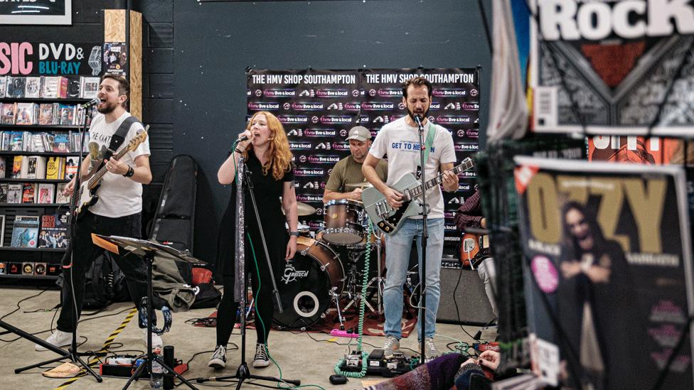 A female-fronted band plays in an HMV store among the DVDs and magazines.