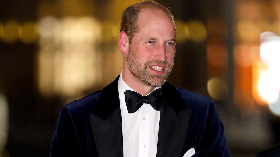 Prince William, the Prince of Wales, smiles as he is photographed arriving at an evening event. He wears a dinner jacket and bow tie with a white shirt. He has close-cropped hair and a short beard and moustache.