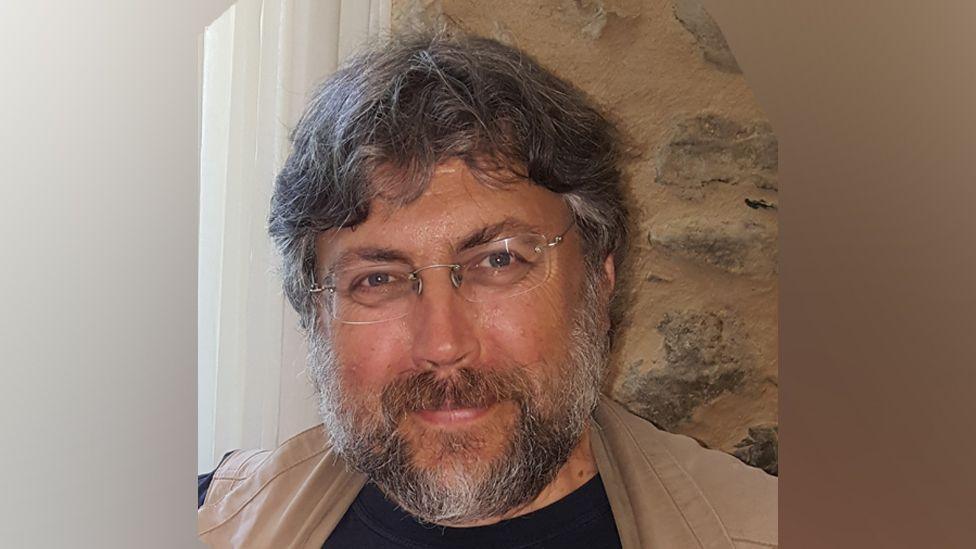 Dr Paul Bahn smiling at the camera. He has black and grey hair and a beard, and he is wearing rimless spectacles and a beige jacket. Behind him is a stone wall.