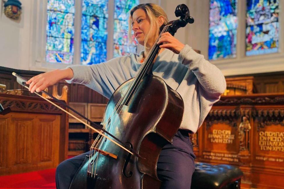 Woman with blonde hair and wearing grey top, playing a dark wood cello, in a chapel setting with stained glass windows in the background