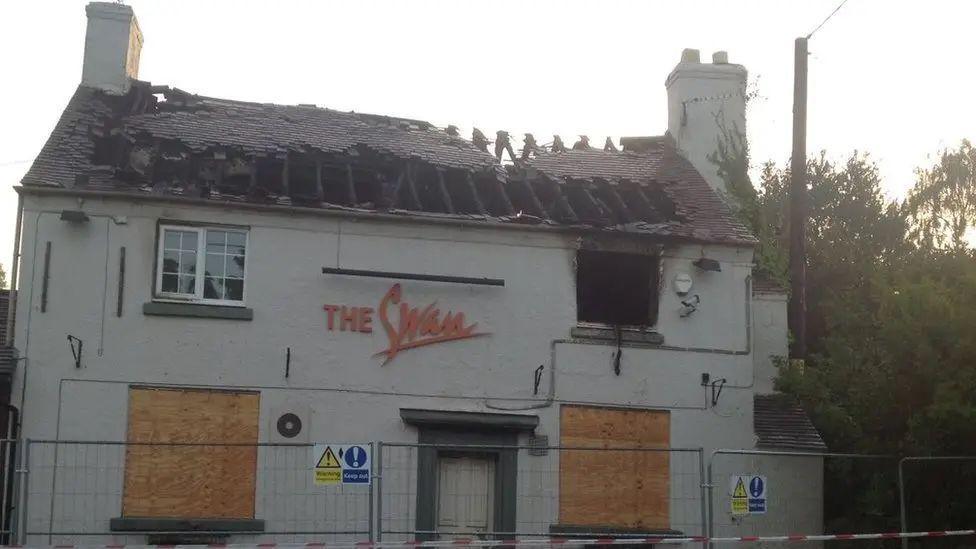 The Swan pub pictured in 2015, it's roof has been destroyed and bottom two windows boarded up. It is a white building with The Swan in red letters