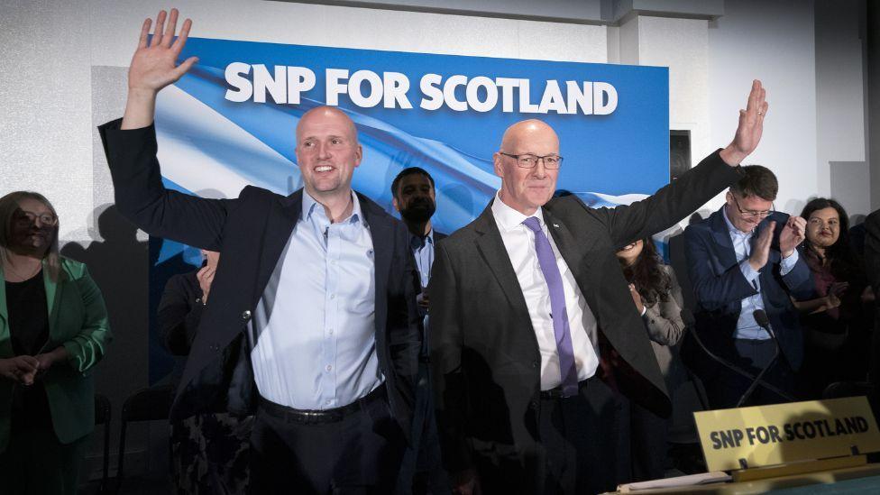 Stephen Flynn and John Swinney, both bald and wearing dark suits, stand beside each other at an SNP campaign event, each with an arm aloft waving in front of clapping colleagues and a blue SNP billboard. 