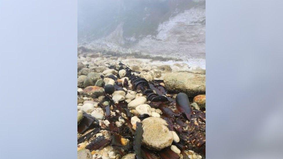 The brown wreckage from the submarine dotted around the large and small rocks of the coastline next to the cliffs. It's misty in the background and the parts are in several different shapes.