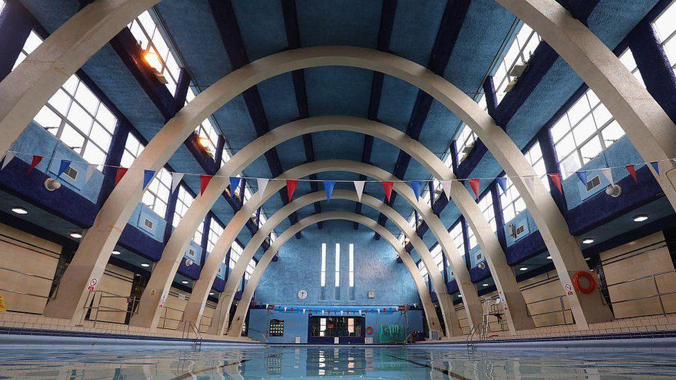 Swimming pool with stone arches above the water.  Large brown doors are visible at the far end. There are high windows and both sides and three long thin windows at the far end.