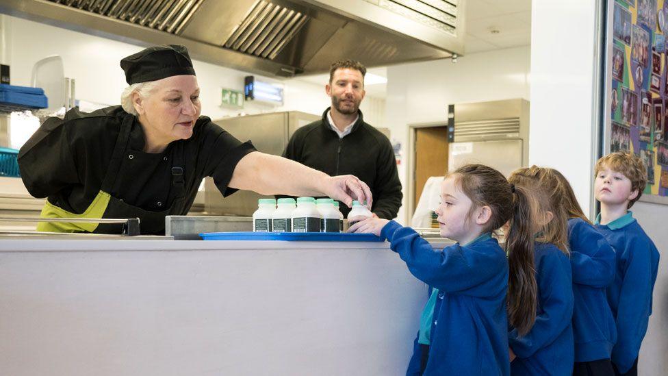 Woman dressed in black chef-style outfit handing milk to a primary school pupil - there are three other pupils in blue jumpers and cardigans behind her.