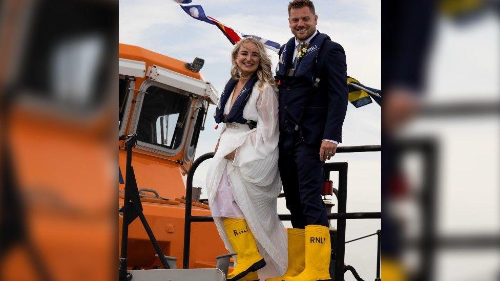 (l-r) Hebe Gregory and Richard Pimm in their wedding attire and wearing yellow RNLI wellies, climbing on board the Severn class lifeboat.
