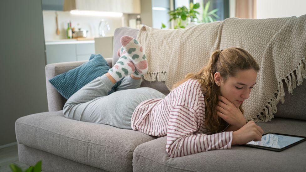 A girl with a pony tail and a stripy top lies on a sofa while using an i-Pad 