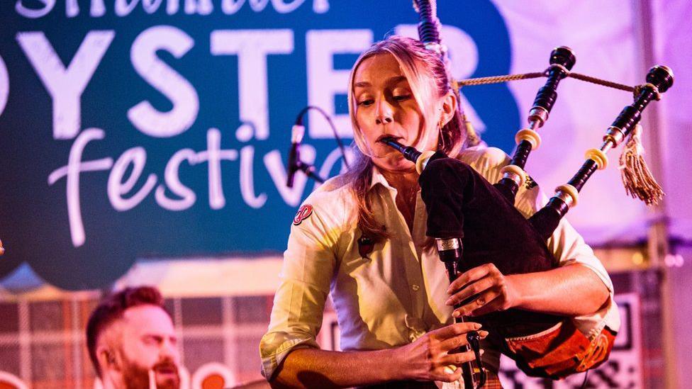 A woman with blonde hair in a yellow blouse plays the bagpipes in front of a Stranraer Oyster Festival sign
