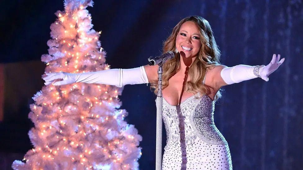 Mariah Carey in front of a microphone in a sparkly dress and long white gloves next to a decorated Christmas tree