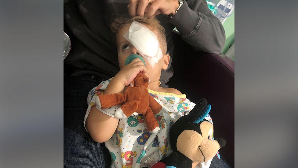 Jace has a white bandage covering one eye and he sits on an adult's knee, cuddling soft toys and wearing a children's colourful hospital gown