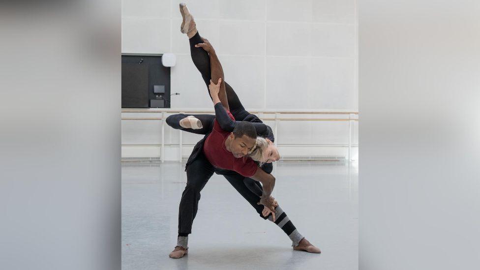 Melissa dancing with with José Alves in a dance rehearsal studio. She is wearing a black unitard and white ballet shoes. José is wearing a red t-shirt and black trousers. He is bent forward holding Melissa on his back. She is grasping his right arm and left leg, he is grasping her left leg and right hand.