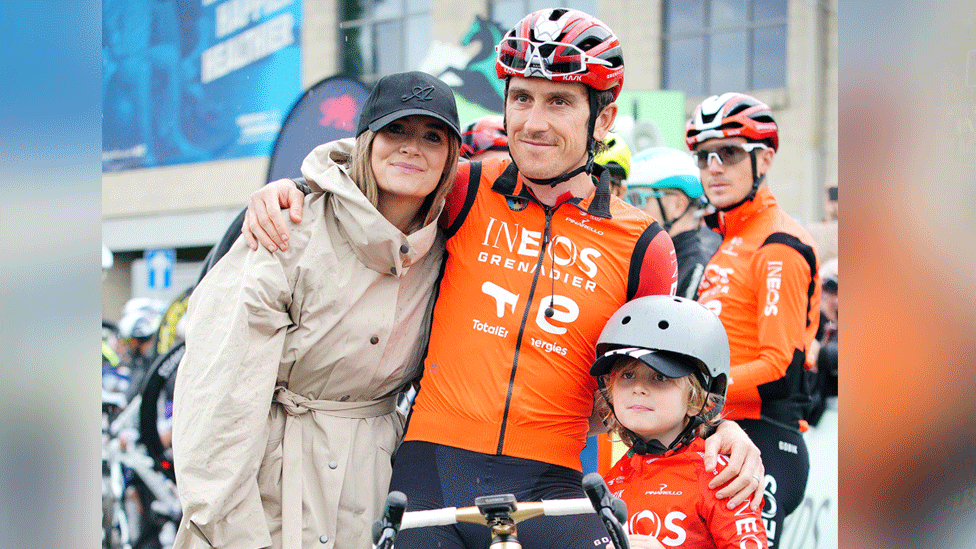 Family photo as the three embrace for a photo. Thomas is in the middle in an orange Ineos Grenadier jacket, and a red helmet on. His son is to his right with the same orange jacket and a white cycle helmet on. His wife is to his left with a black cap and cream rain jacket on. 