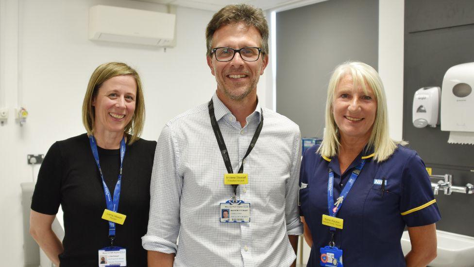 Two female doctors wearing blue landyards and their hospital scrubs. They both have shoulder length blonde hair and are smiling at the camera. They are standing either side of a tall male doctor in the middle, who is wearing a white checked shirt and a black lanyard. They are all standing in the new treatment room