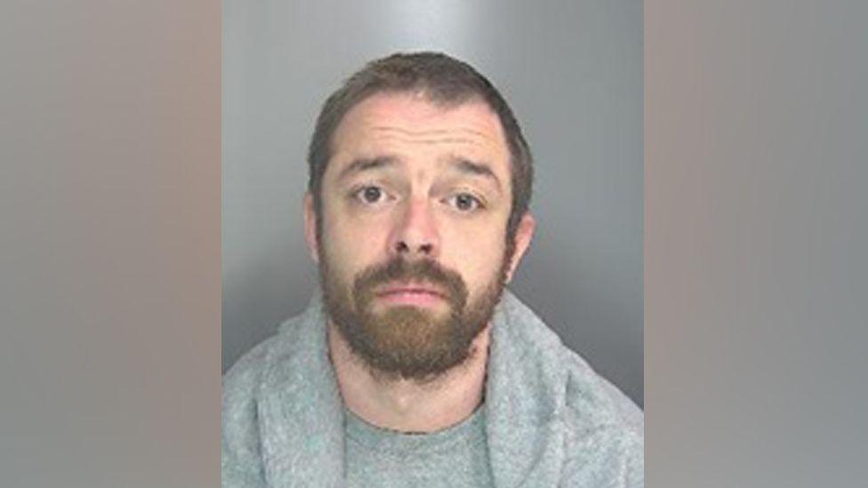 Police custody shot of Adam McDonnell. He is looking at the camera and has brown eyes, short brown hair and a beard and moustache. He is wearing a grey T-shirt and a grey top with a shawl collar
