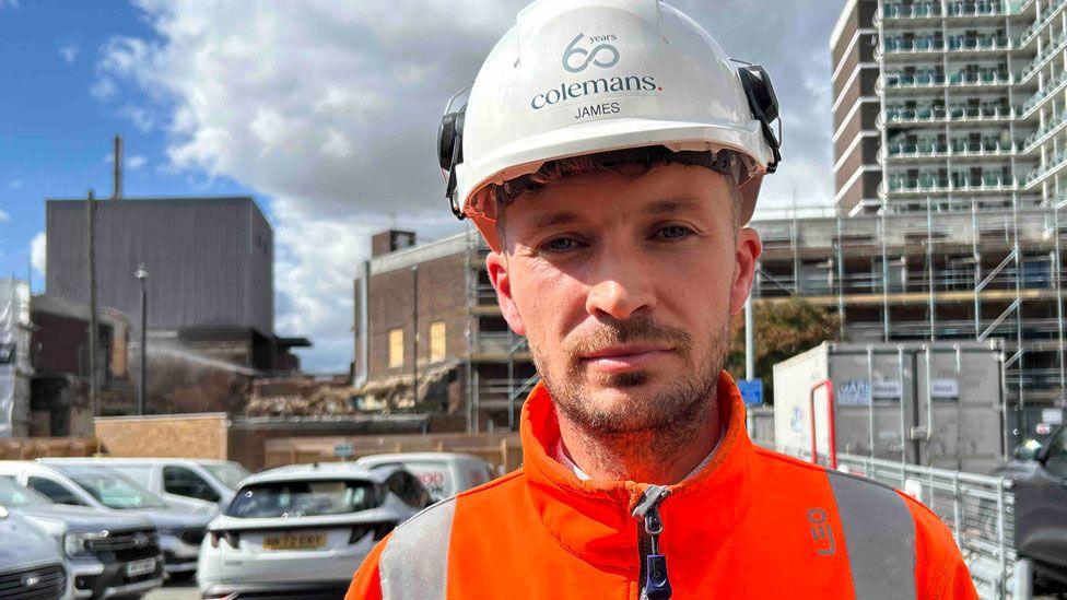 James Doherty who has a short close cropped beard and is wearing a white hard hat with Colemans written on it, and a bright orange hi-vis jacket done up to his neck. Beyond him is a car park with several cars and beyond that are various post-war looking commercial buildings and on the far right a post-war looking block of flats.