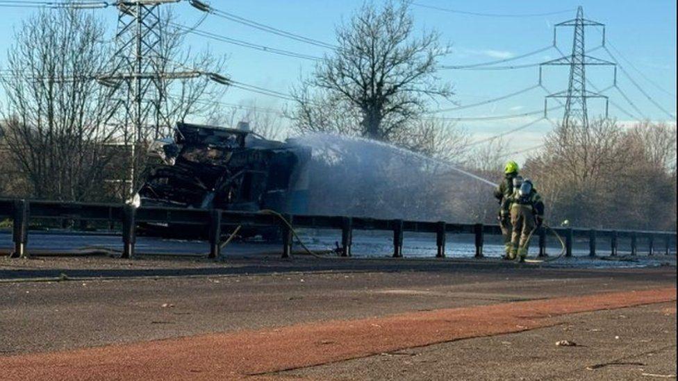 Two firefighters standing behind road barrier with water firing from jets at a blackened van.