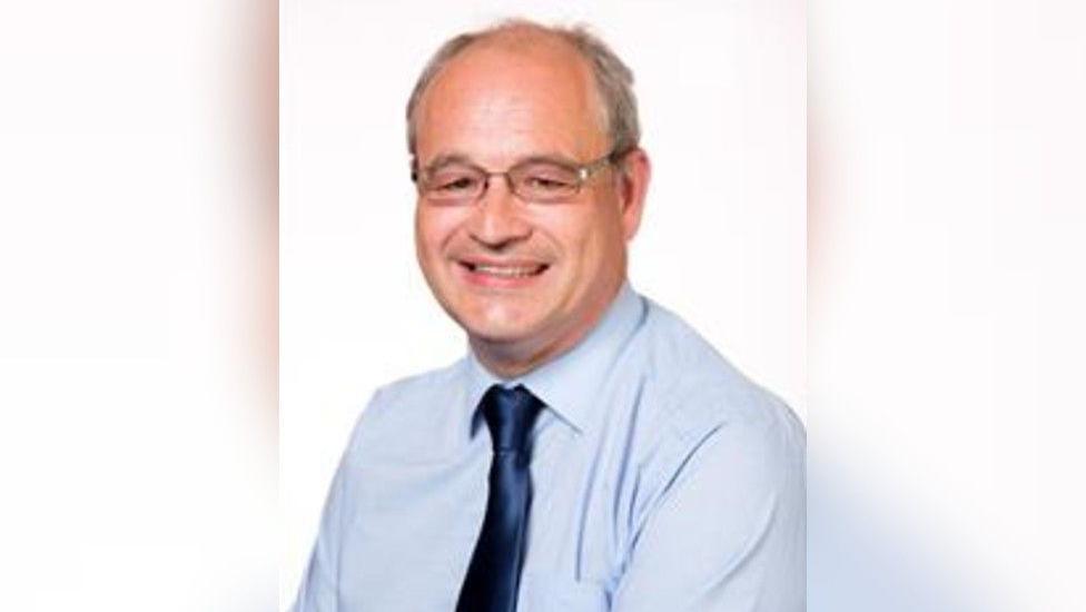 A man with grey hair and glasses smiles at the camera in front of a white background.