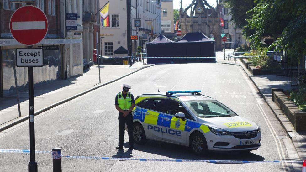 A police officer in front of a police car at the scene of the incident. There is police tape at both ends of a street and a large blue police tent in the distance. 