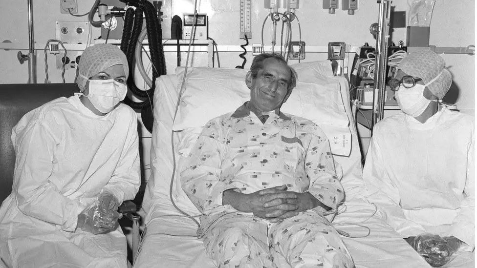 A black and white photo of Keith propped up in a hospital bed, looking at the camera and smiling. He wears a hospital gown or pyjamas, and has various tubes attached. On his left and right sit two women wearing white hospital gowns and plastic gloves. There are various hospital instruments and tubes behind him.