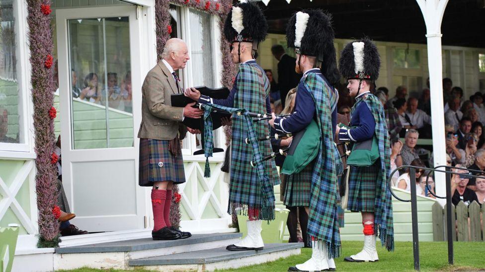 Charles in a brown tartan kilt meets three pipers wearing green tartan and bearskin hats