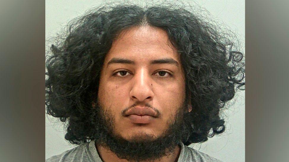 Qusai Abdalla's mugshot. He has a dark beard, brown eyes and shoulder-length, bushy dark hair with a central parting. He is wearing a grey t-shirt and is in front of a plain grey background