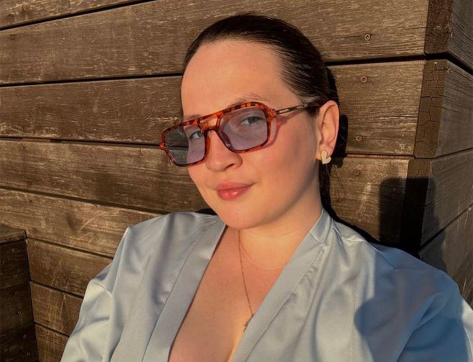 In a selfie taken at sunset, a young woman with brown hair tied back is sitting outside a wooden gite She wears a low-cut grey top, earrings and sunglasses, and is smiling at the camera. 