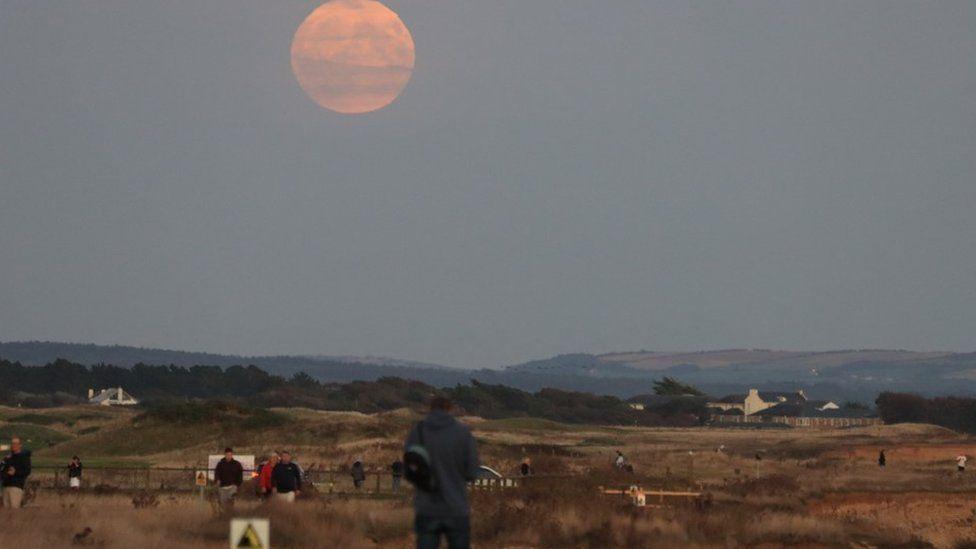 Large orange moon with people standing on heathland watching