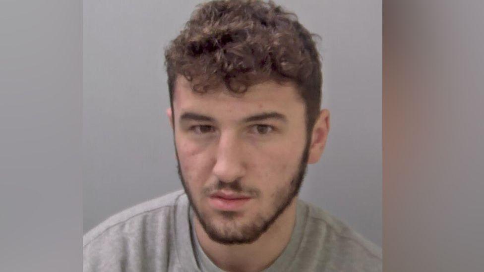 Zack O'Keefe in his custody image, with a curly brown fringe, a slight beard and a grey jumper.