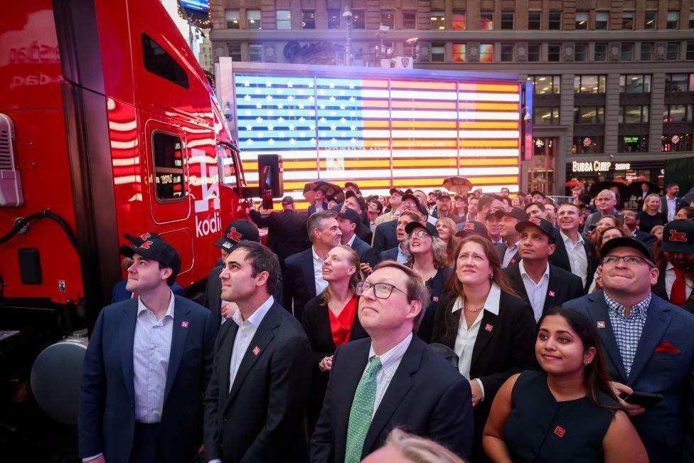 People dressed in suits are in Times Square looking upwards off camera next to a red truck that says Kodiak AI on its side.