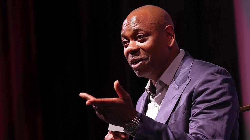 Dave Chappelle speaks at the premiere of "Dave Chappelle: Live In Real Life"  during the 2025 Martha's Vineyard African American Film Festival at Martha's Vineyard Performing Arts Center on August 08, 2025 in Massachusetts.  He is wearing a purple suit and holding a microphone.