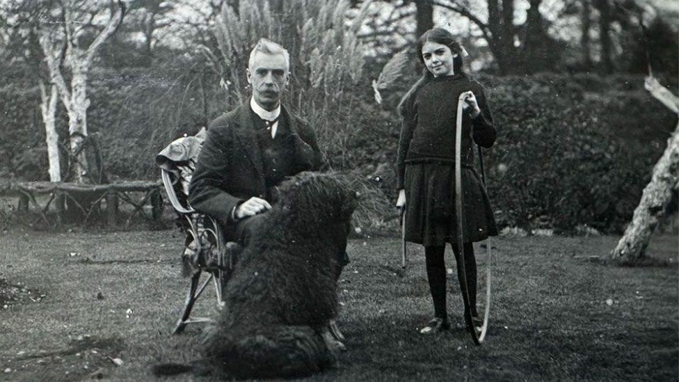 A black and white image of William and Elsa Hall in a garden. He has a high white collar under a dark jacket and waistcoat and is sitting in a garden chair with a large woolly black dog in front of him. She has long dark hair pulled back from her face and is wearing a dark jumper over a dark skirt and is holding a hoop in her left hand.