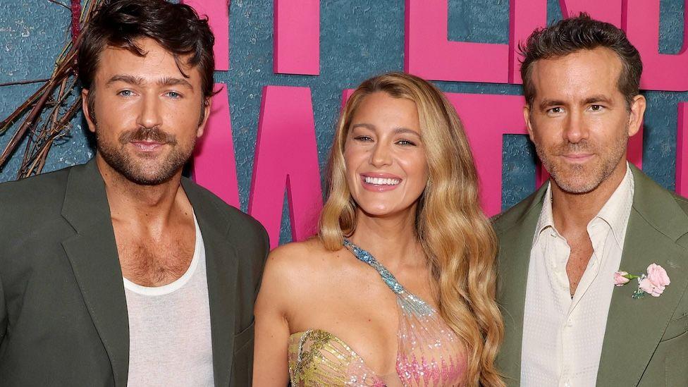 Three people - Brandon Sklenar, Blake Lively and Ryan Reynolds - are dressed in formal attire and posing for a photo. They are stood in front of a large sign that reads "It Ends With Us"