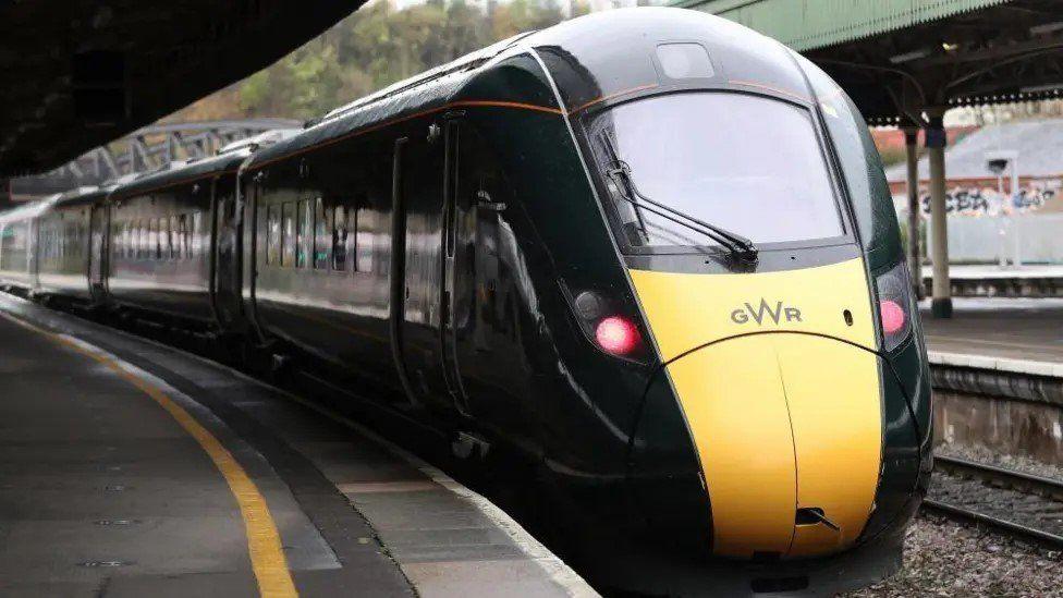 Yellow front of train coming towards the camera with two red lights lit and dark green carriages behind -railway platforms either side.