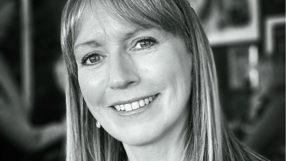 A black and white photo of a woman with long hair. She is smiling at the camera. The background is blurred.
