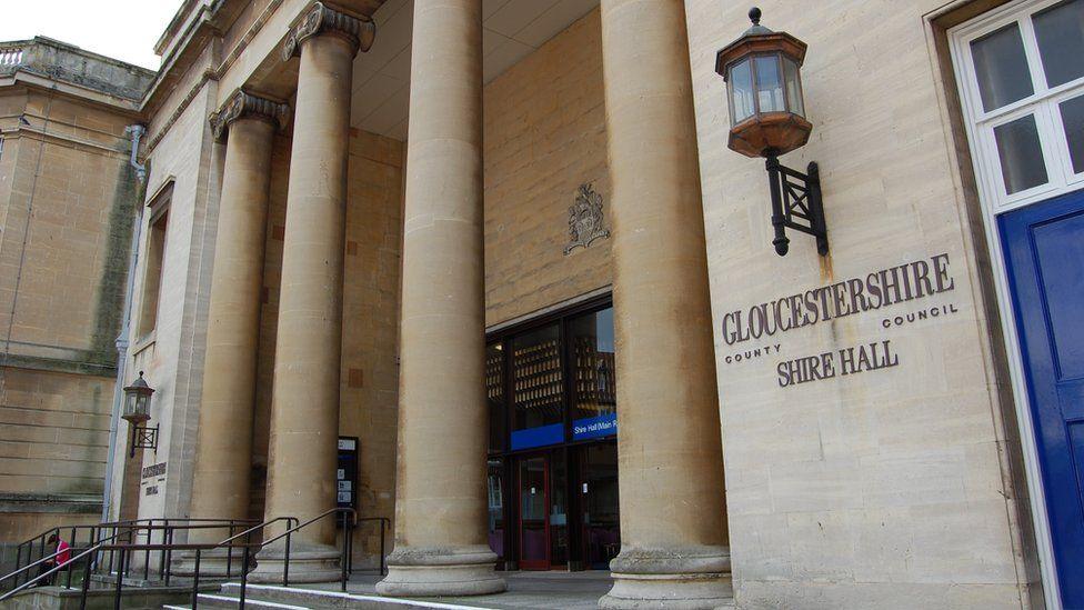 The exterior entrance of Shire Hall in Gloucestershire. It is a large sand-coloured building with formidable stone columns outside. There is a large lantern hanging above a sign which says 'Gloucestershire County Council, Shire Hall'. 