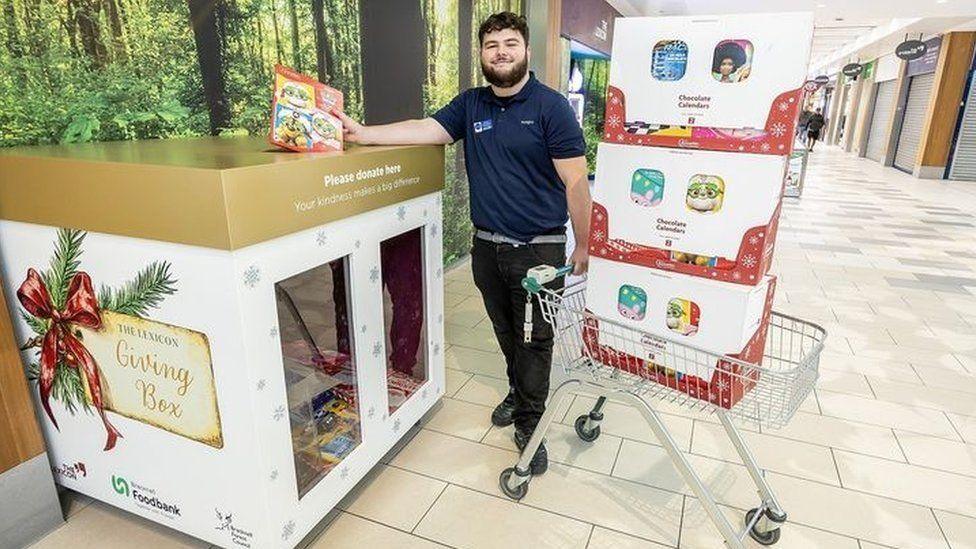 Man standing beside gold and white box posting an advent calendar into it with shopping trolley piled up with boxes.