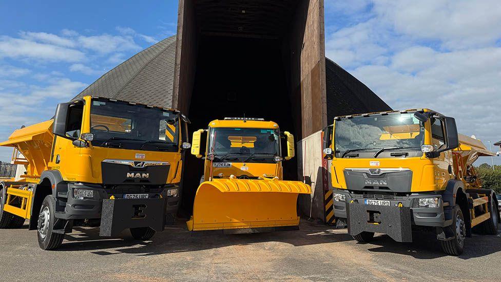 Two yellow and black gritter machines parked either side of a yellow snow plough outside a large domed grit store